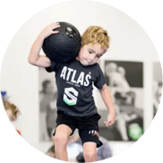 A focused and determined KidStrong elementary student carrying a medicine ball on their shoulder as they navigate through a circuit in a dynamic KidStrong class.