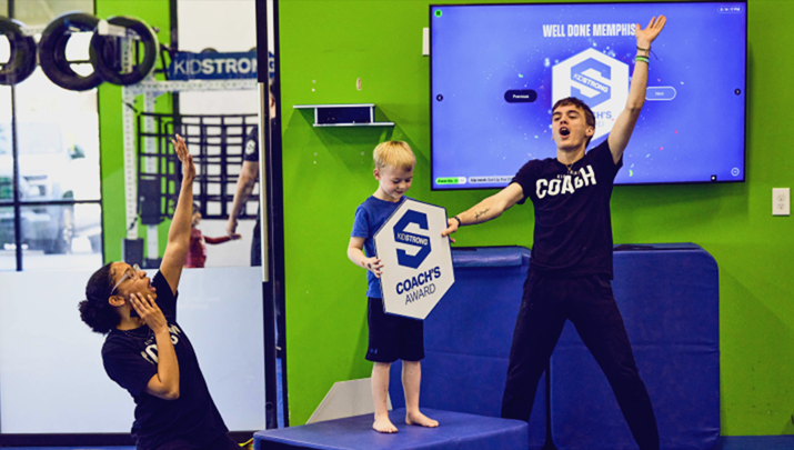 Three focused and determined KidStrong elementary students carrying a medicine ball on their shoulders as they navigate through a circuit in a dynamic KidStrong class.