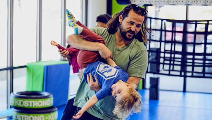 A KidStrong coach pointing towards a screen, teaching a group of enthusiastic elementary KidStrong students about the names of muscles on the human body, fostering an engaging and educational environment.