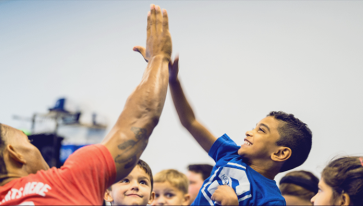 A group of students in the background, with one elementary student in focus wearing a smile of excitement and accomplishment while enthusiastically high-fiving a KidStrong coach.