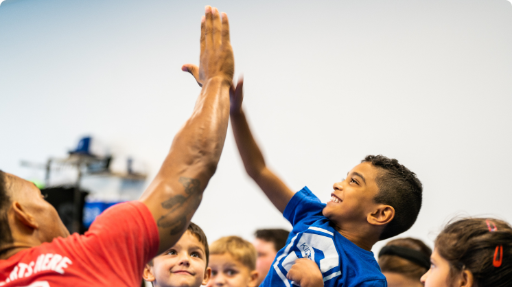 A group of students in the background, with one elementary student in focus wearing a smile of excitement and accomplishment while enthusiastically high-fiving a KidStrong coach.