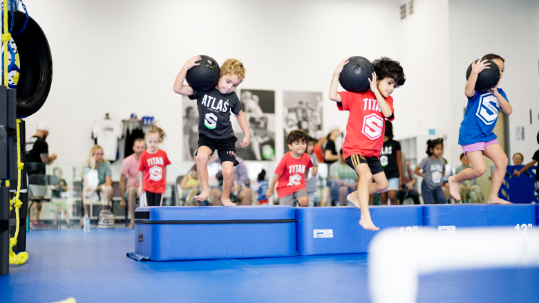 Three focused and determined KidStrong elementary students carrying a medicine ball on their shoulders as they navigate through a circuit in a dynamic KidStrong class.