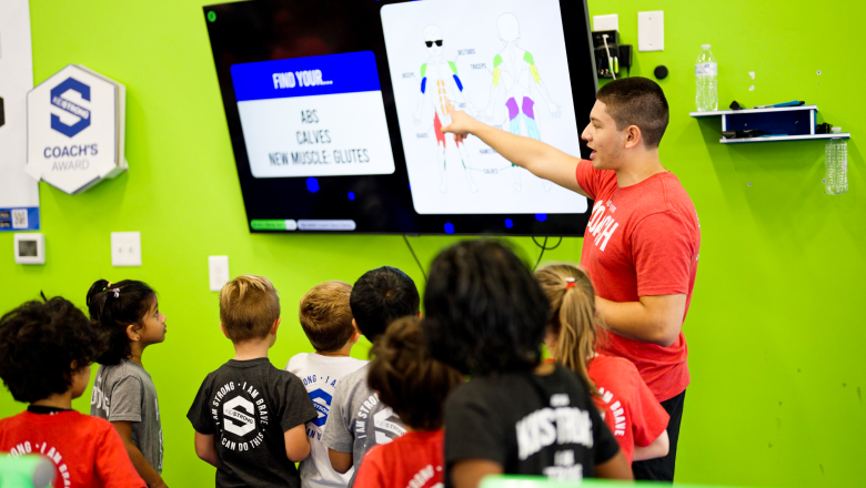 A KidStrong coach pointing towards a screen, teaching a group of enthusiastic elementary KidStrong students about the names of muscles on the human body, fostering an engaging and educational environment.